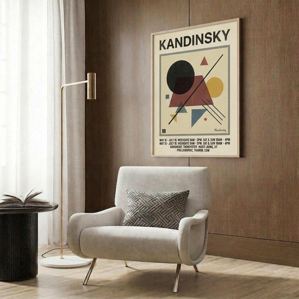 May include: Kandinsky abstract geometric art poster in light wood frame hanging on wood paneled wall above cream upholstered armchair with geometric pillow. Floor lamp with brass finish stands nearby next to dark round side table with open book. Poster features colorful triangles, circles and lines in red, blue, yellow, green and black on cream background.