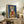 May include: Framed artwork featuring a tabby cat wearing a yellow hooded raincoat and holding a red fish, set against a deep blue background. The print sits on a rustic wooden sideboard alongside books, a small plant, and ceramic vases in a cozy living room setting with neutral walls and brown leather furniture.
