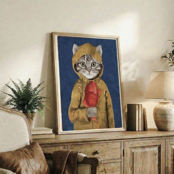 May include: Framed artwork featuring a tabby cat wearing a yellow hooded raincoat and holding a red fish, set against a deep blue background. The print sits on a rustic wooden sideboard alongside books, a small plant, and ceramic vases in a cozy living room setting with neutral walls and brown leather furniture.