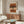 May include: Abstract art print in wooden frame hanging above mid-century walnut sideboard in neutral living room. Artwork features horizontal brushstrokes in terracotta, brown, and cream tones with black dotted pattern down center. Modern curved white chair and glass-top coffee table visible. Natural lighting creates warm shadows on beige walls.