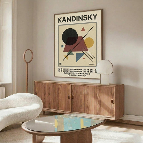 May include: Kandinsky exhibition poster with abstract geometric shapes in black, red, yellow, and blue displayed above a wooden credenza in a modern living room. White curved chair and oval glass coffee table visible in foreground. Poster shows circles, triangles, and intersecting lines in minimalist composition with cream background and black text.