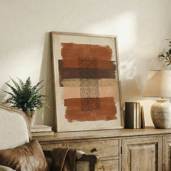 May include: Framed abstract art print with horizontal terracotta, brown, and cream brushstrokes and dotted pattern details, displayed on rustic wood sideboard alongside fern plant, ceramic vase, and stacked books in neutral-toned living room setting.
