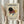 May include: Hands holding a framed abstract geometric art print featuring circles, triangles, and lines in black, burgundy, yellow, and blue on cream background. Person wears beige ruffled sleeves while holding dried pampas grass. Light wooden frame against soft neutral curtains and bright window lighting.