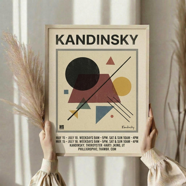 May include: Hands holding a framed abstract geometric art print featuring circles, triangles, and lines in black, burgundy, yellow, and blue on cream background. Person wears beige ruffled sleeves while holding dried pampas grass. Light wooden frame against soft neutral curtains and bright window lighting.