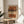 May include: Abstract art print in wooden frame displayed on round wooden side table next to beige upholstered chair. Artwork features horizontal brushstrokes in terracotta, ochre, brown and cream tones with black dotted pattern running vertically through center. Soft natural lighting creates shadows on neutral wall. Green plant leaves visible in corner of minimalist modern living space.