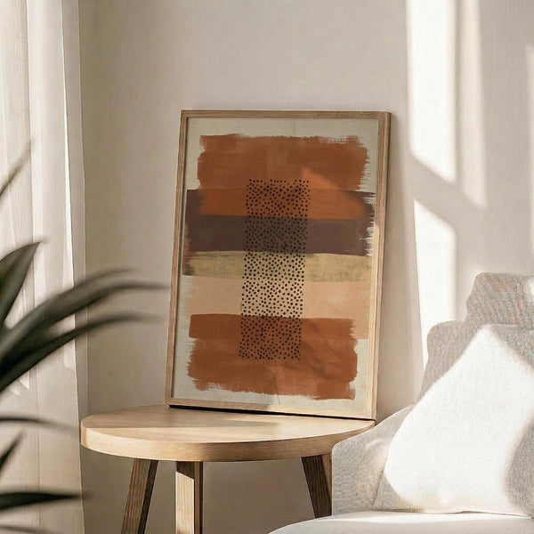 May include: Abstract art print in wooden frame displayed on round wooden side table next to beige upholstered chair. Artwork features horizontal brushstrokes in terracotta, ochre, brown and cream tones with black dotted pattern running vertically through center. Soft natural lighting creates shadows on neutral wall. Green plant leaves visible in corner of minimalist modern living space.