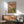 May include: Framed poster print featuring two cartoon ducks in tropical shirts enjoying a picnic with wine, bread, cheese and fruit on a green table against a coral pink background. Displayed above a wooden sideboard with table lamp in a mid-century modern living room with teal upholstered chair and natural lighting casting shadows on white walls.