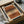May include: Abstract art print in black frame displayed in cardboard shipping box with protective tissue paper. Artwork features horizontal brushstrokes in terracotta, ochre, and cream tones with three dotted stipple patterns overlaid. Warm earth-toned palette with organic painted textures. Frame sits among crumpled white tissue paper with wooden dowels visible nearby.