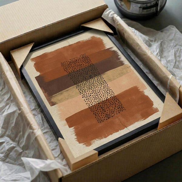 May include: Abstract art print in black frame displayed in cardboard shipping box with protective tissue paper. Artwork features horizontal brushstrokes in terracotta, ochre, and cream tones with three dotted stipple patterns overlaid. Warm earth-toned palette with organic painted textures. Frame sits among crumpled white tissue paper with wooden dowels visible nearby.