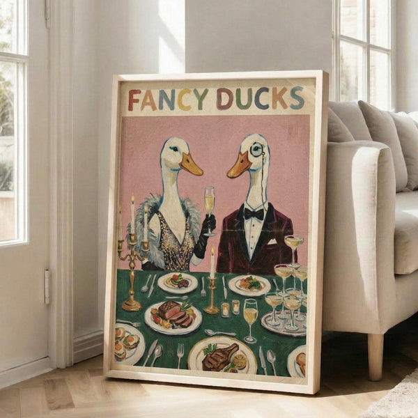 May include: Framed vintage-style poster showing two elegantly dressed ducks at a formal dinner table. The female duck wears a sparkly dress with fur stole, the male duck sports a burgundy tuxedo with bow tie and monocle. They're dining at a green tablecloth-covered table with multiple plates of food, champagne glasses, and candles. The poster has "FANCY DUCKS" text at top in colorful letters against a pink background. Frame leans against white wall near beige couch in bright room.
