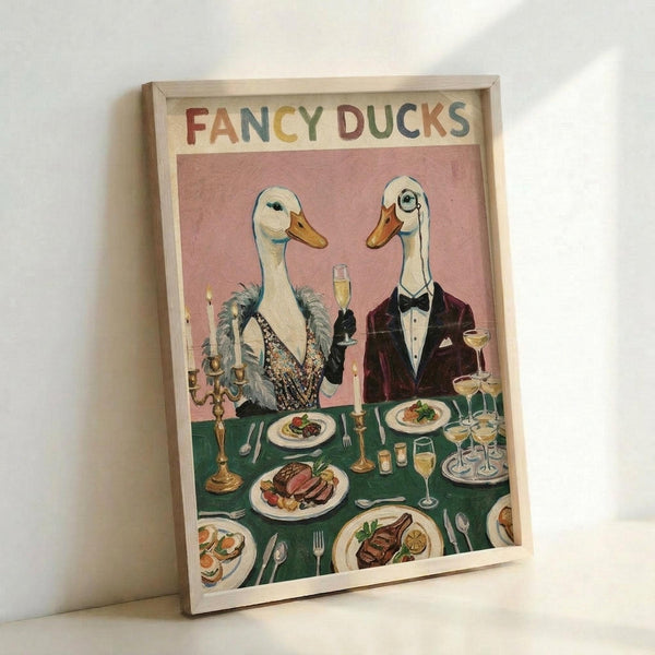 A framed vintage-style illustration showing two anthropomorphic ducks dressed formally at an elegant dinner table. The female duck wears a sequined dress with fur stole, while the male duck sports a burgundy tuxedo with bow tie. They hold champagne glasses above a green tablecloth set with fine dining plates, candelabras, and gourmet food. The artwork has a pink background with "FANCY DUCKS" lettering in colorful text at the top, displayed in a light wood frame leaning against a white wall.