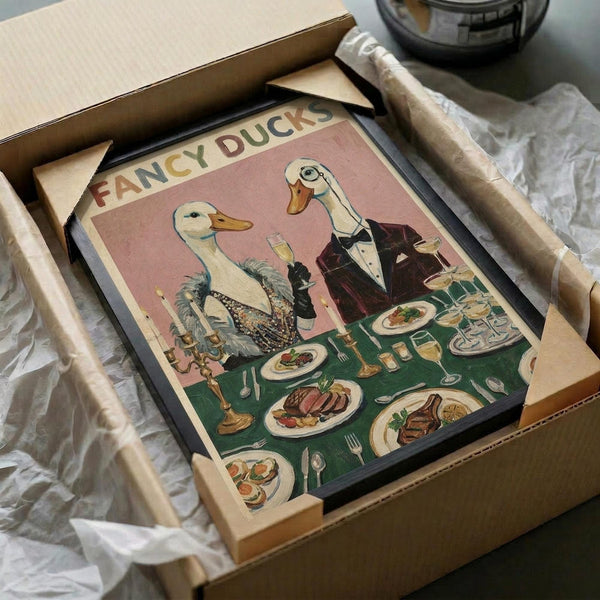 May include: Framed vintage-style poster showing two elegant ducks in formal attire dining at a green tablecloth-covered table. The female duck wears a beaded black dress while the male duck sports a burgundy tuxedo. Multiple plates of gourmet food and wine glasses surround them on the table. The poster has a soft pink background with "FANCY DUCKS" text at the top. The black-framed artwork is nestled in shipping packaging with protective wrapping materials.