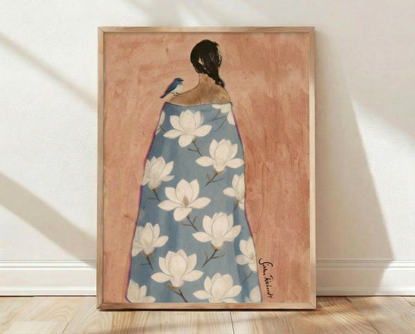 May include: Framed artwork leaning against white wall on wooden floor. The piece shows a woman from behind with dark braided hair and a small blue bird perched on her shoulder. She wears a flowing blue robe decorated with white magnolia flowers. The background has warm peachy-pink tones with a textured, watercolor-like appearance. Natural wooden frame surrounds the artwork. Soft natural lighting creates gentle shadows on the wall.