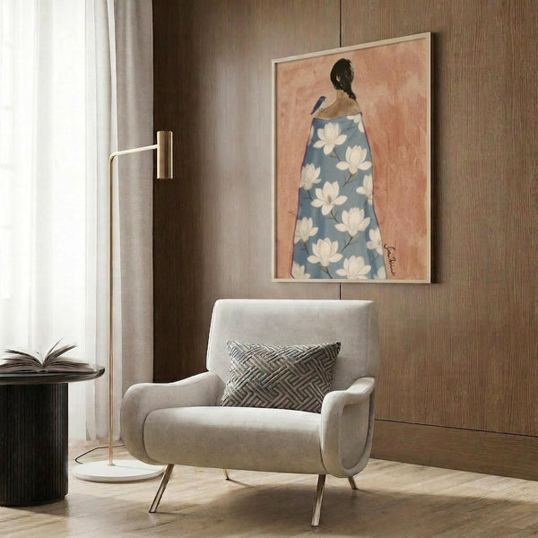 May include: Framed artwork showing a woman from behind wearing a blue robe with white magnolia flowers, set against a warm peach background, displayed on wood paneled wall above a beige upholstered armchair with geometric pillow, brass floor lamp and side table with open book nearby