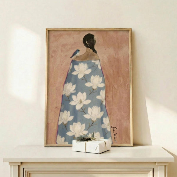 May include: Framed artwork showing a woman from behind with dark hair in a braid, wearing a blue robe decorated with white magnolia flowers, with a small blue bird perched on her shoulder. The painting has a soft pink background and is displayed in a light wood frame on a white wooden shelf alongside a small wrapped gift with greenery.