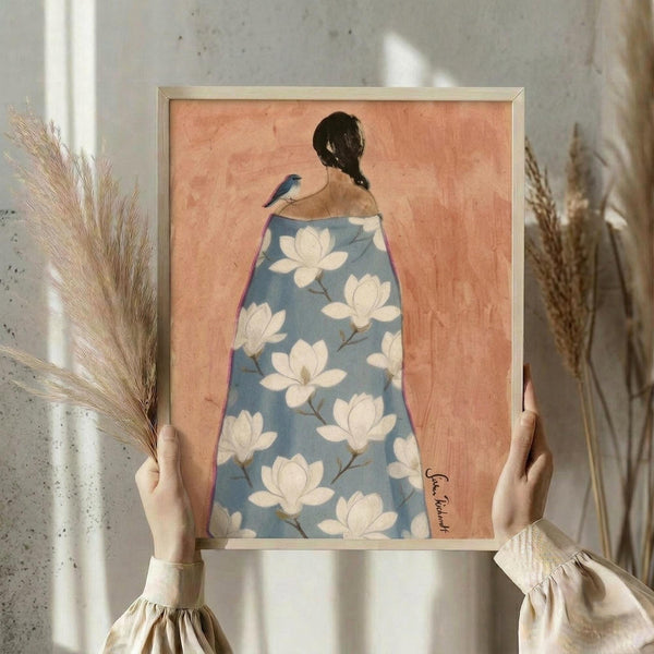 May include: Hands holding framed artwork showing woman from behind with dark braided hair and small blue bird perched on shoulder, wearing flowing robe with white magnolia flowers on soft blue background against peachy coral backdrop. Natural wood frame, neutral home setting with dried pampas grass in background, cream colored sleeves visible.