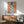 May include: Framed artwork showing a woman from behind wearing a blue robe decorated with white magnolia flowers, displayed above a wooden sideboard with table lamp in a modern living room setting with sage green armchair and natural lighting creating soft shadows on white walls.