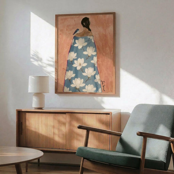 May include: Framed artwork showing a woman from behind wearing a blue robe decorated with white magnolia flowers, displayed above a wooden sideboard with table lamp in a modern living room setting with sage green armchair and natural lighting creating soft shadows on white walls.