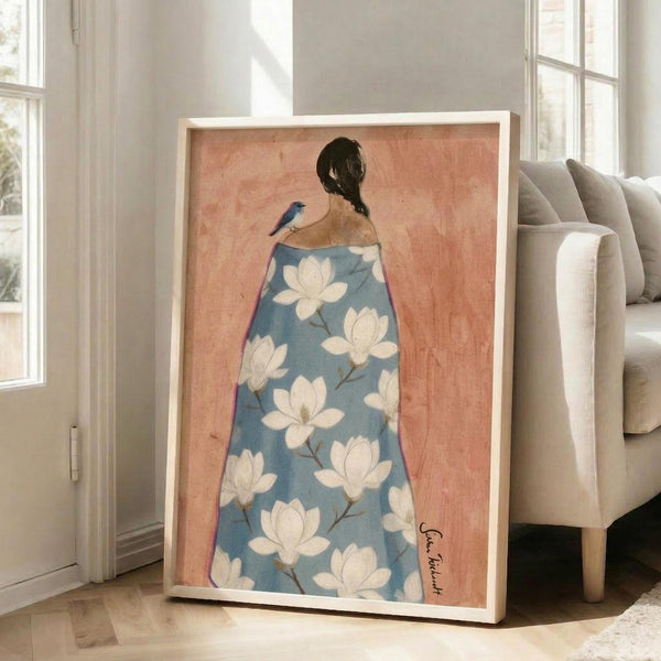 May include: Framed art print leaning against white wall next to beige sofa showing woman from behind wearing blue robe with white magnolia flower pattern, small bird perched on her shoulder, coral pink background, natural lighting from windows, light wood frame on hardwood floor