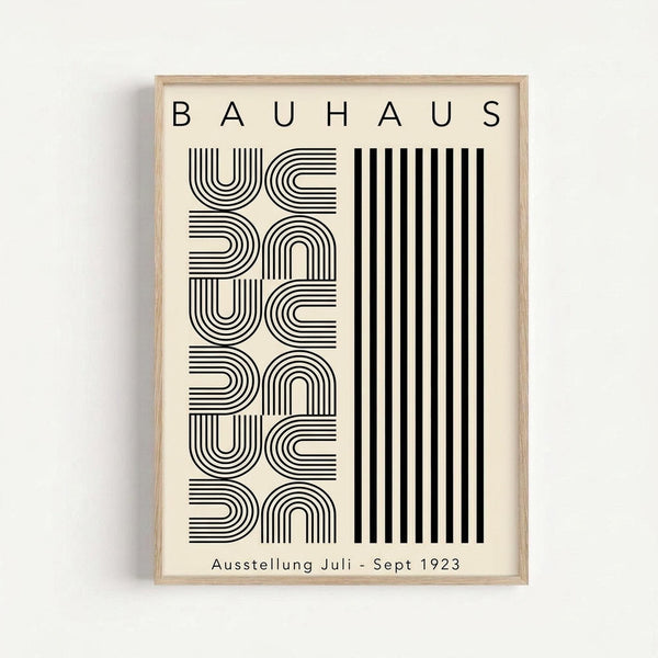 A framed poster with cream background featuring black text reading "BAUHAUS" at top, geometric letterforms spelling "BAUHAUS" in curved and straight lines on the left side, vertical black stripes on the right side, and "Ausstellung Juli - Sept 1923" at bottom. The minimalist design uses only black ink on cream paper, displayed in a light wood frame against a white wall.