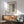 May include: Modern living room mockup showing framed Bauhaus exhibition poster on white wall above wooden sideboard. Poster features black geometric typography and stripes on cream background in natural wood frame. Mid-century furniture includes sage green upholstered armchair with wooden arms, round coffee table, and ceramic table lamp. Bright natural sunlight creates dramatic shadows across the minimalist interior space.