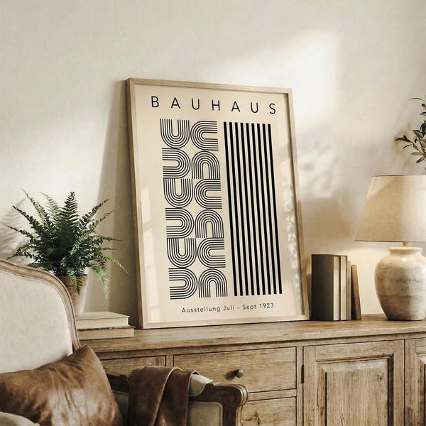 May include: Framed Bauhaus exhibition poster with black geometric typography and stripes on cream background, displayed on rustic wood sideboard alongside fern plant, ceramic vase, and stacked books in neutral-toned living room setting with upholstered chair