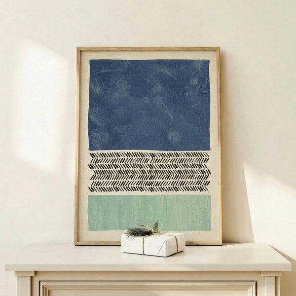 May include: Framed abstract print with three horizontal sections - textured navy blue at top, black and white herringbone pattern in middle, and sage green at bottom. Light wood frame sits on pale wooden console table against cream wall. Small white wrapped gift with greenery sprig placed in front of artwork.