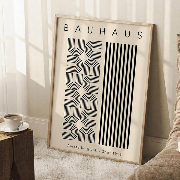 May include: Framed Bauhaus-style poster with black geometric lettering on cream background, leaning against white wall corner. Wooden frame, displayed on white textured rug beside wooden side table with white coffee cup. Natural lighting creates soft shadows on minimalist interior setting.