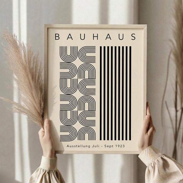 May include: Person holding framed Bauhaus exhibition poster in natural wood frame. Black geometric typography on cream background featuring curved and straight line patterns. Dried pampas grass visible on left side. Neutral beige clothing and soft natural lighting create minimalist aesthetic. Poster shows vintage German exhibition text from 1923.