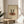 May include: Framed poster showing two silhouetted figures dancing on a red vinyl record, displayed on a round wooden side table next to a beige upholstered chair. Natural sunlight streams across the neutral-toned room, casting soft shadows. The artwork features a cream background with text reading "1970-1980" and "record danar love you" beneath the dancing couple illustration.