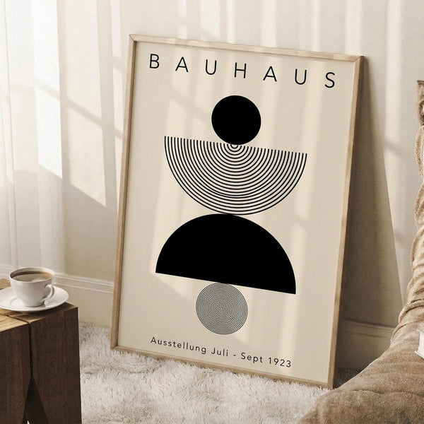May include: Framed Bauhaus poster with geometric black shapes and concentric line patterns on cream background, leaning against white wall corner. Scene shows wooden side table with white coffee cup and saucer, beige knit throw blanket, and white textured rug. Natural window light creates soft shadows across the minimalist room setting.