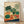 May include: Framed botanical illustration print leaning against white wall on light wood floor. Artwork features various vegetables including leafy greens, carrots, tomatoes, cabbage, radishes, and herbs in green, red, and orange colors against cream background with decorative blue and orange wave elements at bottom. Natural wood frame with simple design.