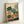 Framed botanical illustration print featuring various vegetables including leafy greens, carrots with orange roots, white turnip, red tomatoes, radishes, cabbages, and tall herbs arranged on a cream background with orange and navy blue brushstroke elements at the bottom. The artwork is displayed in a light wood frame leaning against a white wall.