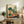 May include: Framed vegetable garden illustration artwork displayed on rustic wooden console table alongside fern plant, books, and ceramic vases. The print features various vegetables like cabbage, tomatoes, radishes, and leafy greens in green, orange, and red colors against cream background with blue water element at bottom. Natural wood frame complements the earthy home decor setting with beige chair and neutral wall.
