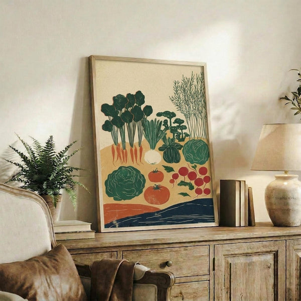 May include: Framed vegetable garden illustration artwork displayed on rustic wooden console table alongside fern plant, books, and ceramic vases. The print features various vegetables like cabbage, tomatoes, radishes, and leafy greens in green, orange, and red colors against cream background with blue water element at bottom. Natural wood frame complements the earthy home decor setting with beige chair and neutral wall.