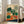 May include: Framed botanical poster featuring illustrated vegetables in green, orange, and red colors against cream background with blue accent at bottom. Shows carrots, cabbages, tomatoes, radishes, and leafy greens in vintage-inspired flat design style. Light wood frame leaning against white wall next to beige upholstered chair in bright, airy living room with large windows.