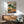 May include: A botanical vegetable garden poster in wooden frame hanging above mid-century modern credenza with table lamp. The artwork features illustrated vegetables including carrots with green tops, tomatoes, cabbages, mushrooms, and leafy greens in earth tones against cream background. Green upholstered armchair with wooden arms sits beside round coffee table in bright sunlit room with white walls.