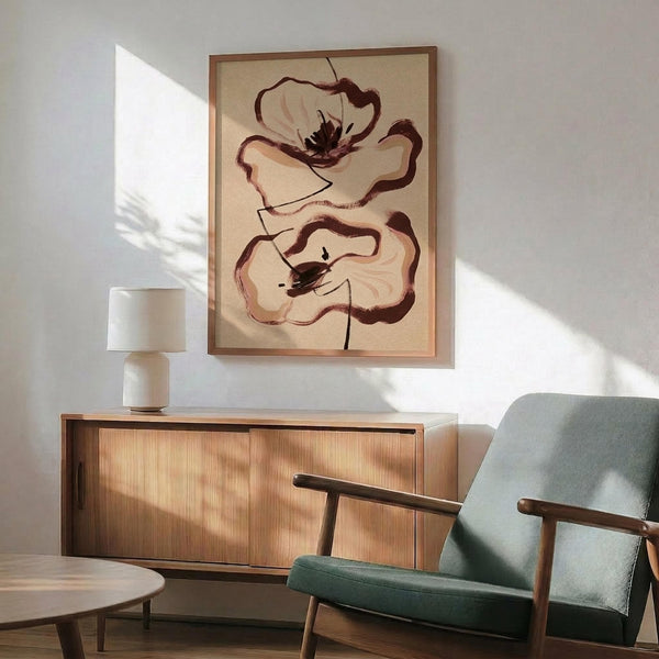 May include: Abstract floral artwork featuring two stylized poppies in earthy brown and cream tones, displayed in a thin wooden frame above a mid-century modern wooden credenza. Scene shows natural sunlight streaming across white walls, with a sage green upholstered armchair, small ceramic table lamp, and round wood coffee table creating a warm, contemporary living room setting.