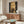 May include: Modern living room displaying a vertical astronomical poster featuring four golden full moons stacked against a dark starry sky background. The artwork hangs above a mid-century walnut credenza with sliding doors, flanked by copper floor lamp and white table lamp. Scene includes cream curved chair and oval glass coffee table with colorful abstract art reflection.