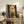 May include: Framed celestial artwork showing four golden moons vertically stacked against a dark starry sky background, displayed on a rustic wooden sideboard alongside potted ferns, ceramic vases, and books in a warm-toned living room setting with cream chair and neutral walls.
