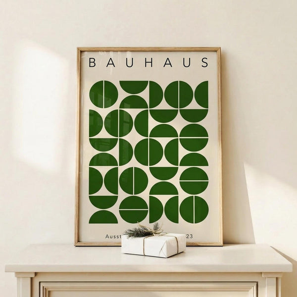 May include: Framed Bauhaus-style poster with geometric green circles and semicircles arranged in a grid pattern on cream background, displayed on light wood surface beside small wrapped gift with twine and dried sprig, against neutral wall with soft natural lighting.