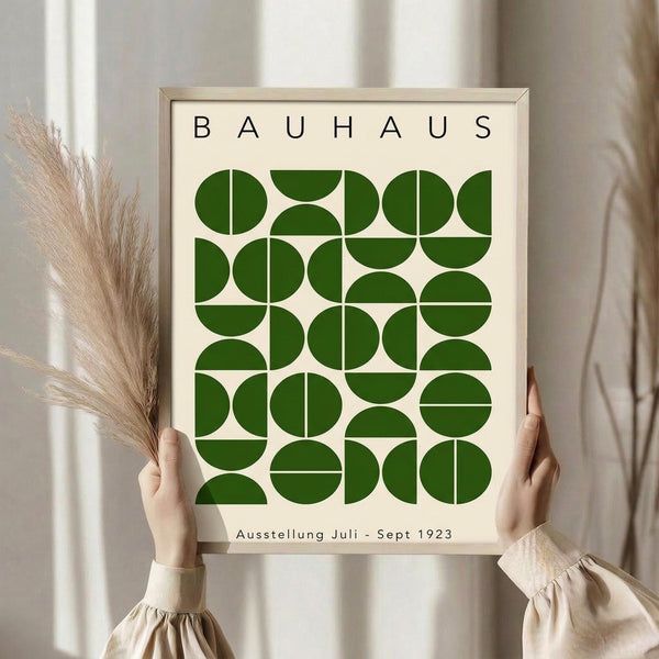 May include: Person in beige ruffled sleeve top holding framed geometric poster with green semicircles and quarter-circles arranged in grid pattern on cream background, "BAUHAUS" text at top and "Ausstellung Juli - Sept 1923" at bottom, dried pampas grass visible to left, soft neutral curtains in background