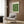 May include: Framed Bauhaus poster with green geometric circles on cream background hanging on wood panel wall above beige mid-century modern armchair with geometric pillow. Modern brass floor lamp and dark round side table with open book visible. Neutral tones throughout minimalist living room setting with natural lighting from window.