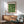 May include: Framed Bauhaus poster with green geometric circles and semicircles pattern hanging on white wall above wooden sideboard. Mid-century modern room setting with sage green upholstered armchair, round coffee table, and ceramic table lamp. Natural sunlight creates shadows across the scene. Poster features cream background with forest green circular shapes arranged in grid pattern.