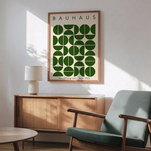 May include: Framed Bauhaus poster with green geometric circles and semicircles pattern hanging on white wall above wooden sideboard. Mid-century modern room setting with sage green upholstered armchair, round coffee table, and ceramic table lamp. Natural sunlight creates shadows across the scene. Poster features cream background with forest green circular shapes arranged in grid pattern.