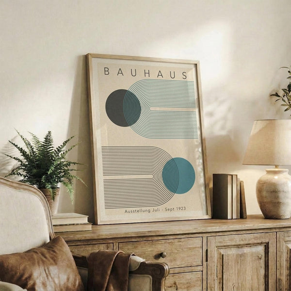 May include: Framed Bauhaus-style poster with geometric design featuring two semi-circular forms in dark green and teal blue with concentric line patterns on cream background, displayed on rustic wooden sideboard alongside books, ceramic vase, and fern plant in modern living room setting with beige chair and neutral decor