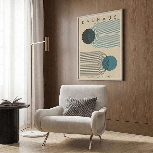 May include: Bauhaus-inspired poster with geometric design featuring curved lines and blue circles on cream background, displayed above modern beige armchair with patterned pillow. Room setting shows wood paneled walls, brass floor lamp, and side table with open book. Minimalist mid-century modern interior styling with warm neutral tones and natural lighting from window.