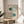 May include: Framed Bauhaus exhibition poster with geometric design featuring two curved linear patterns in teal and sage green with solid circles, displayed on round wooden side table next to beige upholstered chair. Soft natural lighting creates shadows on cream walls, with plant leaves visible in corner of minimalist room setting.