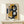 May include: Abstract geometric art print in natural wood frame displaying overlapping shapes in navy blue, mustard yellow, gray, and cream tones with bold black curved forms. Framed artwork leans against white wall on light wood surface next to small white wrapped gift with evergreen sprig, creating modern minimalist home decor display.