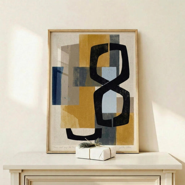 May include: Abstract geometric art print in natural wood frame displaying overlapping shapes in navy blue, mustard yellow, gray, and cream tones with bold black curved forms. Framed artwork leans against white wall on light wood surface next to small white wrapped gift with evergreen sprig, creating modern minimalist home decor display.